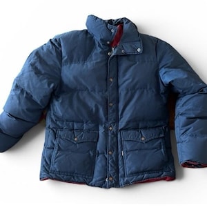 May include: A navy blue puffer jacket featuring a high collar, a front zipper closure with a snap-button placket, and two large front pockets with flap closures. The interior lining is a contrasting dark red color visible at the collar and hem.