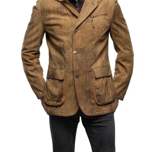 May include: A brown leather blazer with a textured finish. The jacket features a notched lapel, two front pockets, a chest pocket with a zipper, and button closures. The sleeves have elbow patches. The model is wearing dark gray pants.