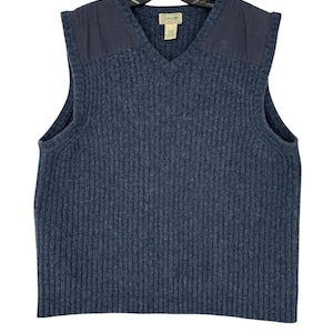 Może przedstawiać: Niebieska kamizelka z dzianiny żeberkowej z dekoltem w serek i naszywkami na ramionach. Kamizelka ma metkę wewnątrz kołnierza. Kamizelka wisi na czarnym wieszaku. Kamizelka jest w rozmiarze M.