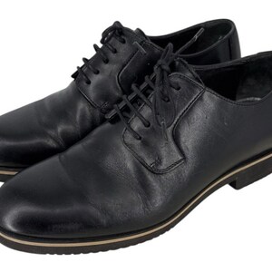 May include: A pair of black leather dress shoes with a lace-up design. The shoes have a rounded toe and a low heel with a tan stripe around the sole. The shoes are on a white background.