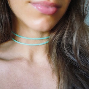 Op de afbeelding: Een close-up van de nek en schouders van een persoon, met twee delicate, licht turkooizen chokers. De chokers zijn dun en zitten dicht op de nek. De persoon heeft lang, golvend bruin haar en draagt de chokers.