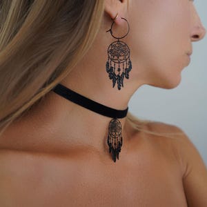 May include: Black dreamcatcher earrings and a matching choker necklace. The earrings are hoop style with a detailed dreamcatcher design, including a floral center and dangling feathers. The choker is a simple black velvet band with a small dreamcatcher pendant.