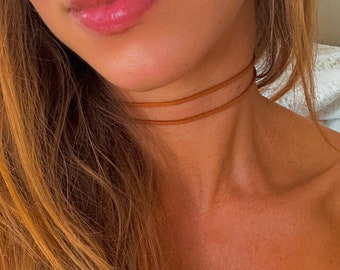 May include: A close-up of a person's neck and face, showcasing a delicate, double-strand brown choker necklace. The skin tone is fair, with long, light brown hair cascading down. The lips are glossy and pink.