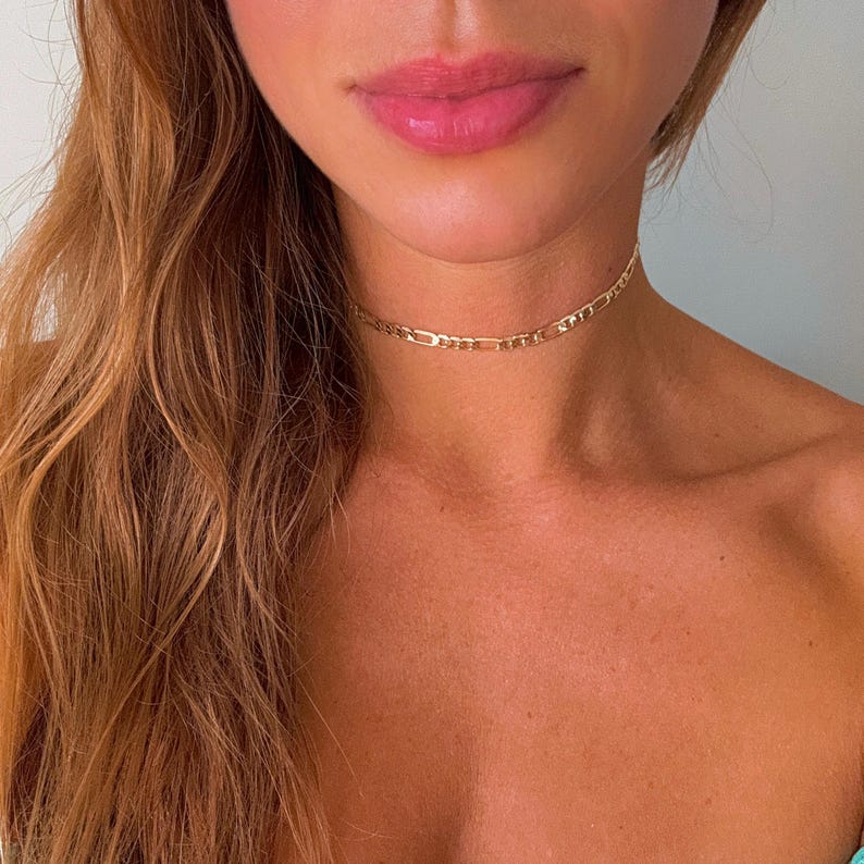 May include: A gold chain choker necklace on a person with long brown hair.