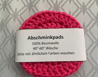 May include: A round, hot pink crocheted pad with a white label. The label reads "Abschminkpads", "100% Baumwolle", "40°-60° Wäsche", and "bitte mit ähnlichen Farben waschen". The pad is made of cotton.
