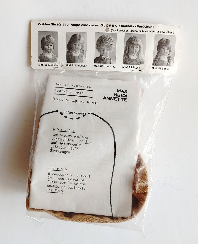 May include: A vintage doll pattern by Max Heidi Annette, packaged in a clear plastic bag. The pattern includes instructions in German and French, with a diagram of the doll's body. The package also features images of doll wigs.
