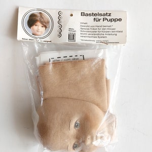 May include: A doll-making kit in a clear plastic bag. The kit includes a doll head with painted features, fabric, and instructions. The packaging has text in German, including "Bastelsatz f&uuml;r Puppe."