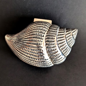 May include: A silver-coloured seashell-shaped decorative object. The shell has detailed ridges and a textured surface, with a small rectangular piece attached at the top. The object is set against a dark background.