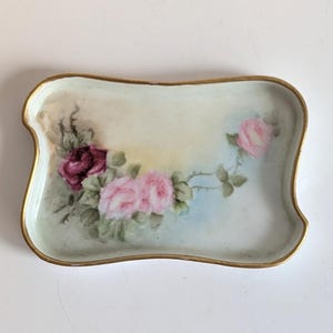 May include: Rectangular porcelain tray with a gold rim and a hand-painted floral design. The tray features pink and burgundy roses with green leaves against a soft, pastel background. A decorative piece for display or serving.