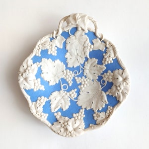 May include: A bright blue ceramic dish with a scalloped edge and a raised white relief pattern featuring detailed grape clusters and vine leaves. The central area is textured, and the decorative border includes a sculpted vine handle at the top.