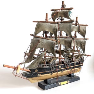 May include: A detailed wooden model ship with weathered grey sails, black hull, and tan accents. The model features multiple masts, rigging, and a decorative base. The ship is a classic nautical decorative item.