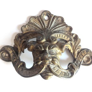 May include: An antique brass drawer pull with an ornate design, featuring a stylized face with a mustache and decorative flourishes. The hardware has a distressed finish and a small hole for mounting. The design is reminiscent of classical or Renaissance styles.