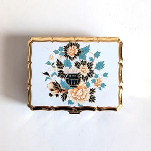 May include: Rectangular trinket box with a white background and a gold-colored scalloped edge. The box features a floral design with peach, black, and teal flowers and leaves, centered around a black vase.