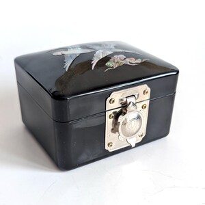 May include: A black lacquered box with a rectangular shape. The lid features an inlaid design of a bird in flight, with silver and iridescent accents. The box has a silver-toned clasp and hinges.