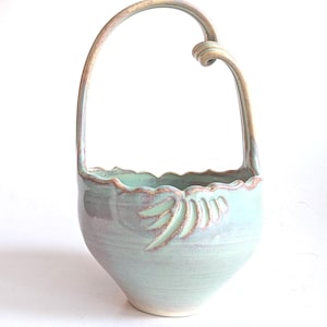 Könnte beinhalten: Ein handgefertigter Keramikkorb mit gebogenem Griff. Der Korb ist ein weiches, gedämpftes Grün mit einem gewellten Rand und einem Blattdesign. Der Griff hat eine hellbraune Farbe.