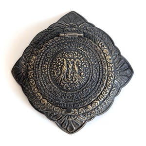 May include: Ornate, antique-style metal box with a hinged lid. The box features intricate embossed patterns and a central medallion with two bird figures. The color scheme is a mix of dark gray and gold, creating a vintage aesthetic.