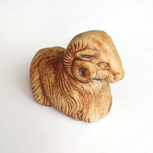 May include: A ceramic ram figurine in shades of tan and brown. The ram is depicted in a seated position with detailed fur and curled horns. The sculpture has a rustic, handcrafted appearance, suitable for home decor.