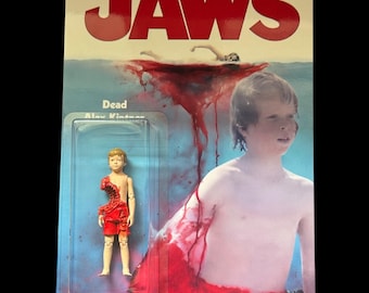 Może przedstawiać: Figurka akcji Jaws na karcie. Karta zawiera tytuł filmu w czerwonych literach, scenę pływaka w wodzie i grafikę krwi. Figurka akcji przedstawia chłopca w czerwonym stroju z ugryzieniem rekina.