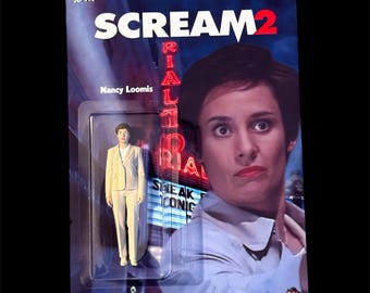 May include: A Scream 2 action figure of Nancy Loomis in a clear plastic package. The packaging features a blue background with the movie title and a photo of the actress. The figure is wearing a cream-colored suit.
