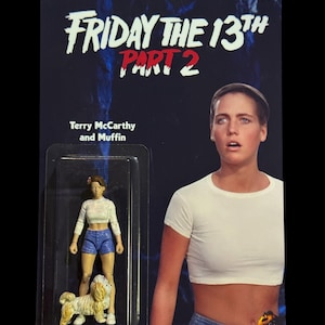 May include: A vintage action figure card for "Friday the 13th Part 2" featuring a figure of Terry McCarthy and her dog Muffin. The card has a dark blue background with the movie title in white and red lettering. The figure is in a clear plastic case.
