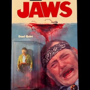 Może przedstawiać: Opakowanie figurki akcji w stylu vintage z tytułem filmu "JAWS" w czerwonych literach. Opakowanie zawiera figurkę "Dead Quint" i graficzny obraz sceny z filmu. Tło przedstawia krwawą scenę oceanu.