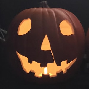 Może przedstawiać: Wyrzeźbiona dynia z zapaloną świecą w środku, tworząca efekt świecenia. Dynia ma tradycyjną twarz jack-o'-lantern z tr&oacute;jkątnymi oczami i poszarpanymi ustami. Pomarańczowa dynia jest na ciemnym tle.