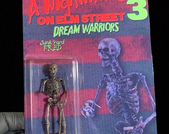 Op de afbeelding: Een Nightmare on Elm Street 3: Dream Warriors actionfiguur van Freddy Krueger als skelet met een schop. De figuur is verpakt op een rood-zwart gestreepte kaart met de titel van de film en de naam van het personage.