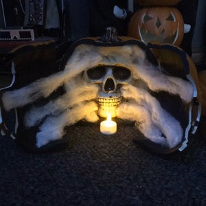 Może przedstawiać: Dekoracja na Halloween przedstawiająca czaszkę i zapaloną świecę wewnątrz dyni. Dynia jest pomarańczowa i czarna, z białą watą wokół czaszki. Mała, zapalona świeca znajduje się przed czaszką.