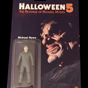 Może przedstawiać: Figurka akcji Halloween 5: The Revenge of Michael Myers w oryginalnym opakowaniu. Karta zawiera dużą, ciemną ilustrację twarzy oraz tytuł filmu. Mała figurka Michaela Myersa w szarym kombinezonie jest zamknięta w plastikowym opakowaniu.