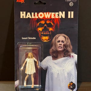 May include: A plastic action figure of a woman in a white hospital gown with a bandage on her leg. The figure is packaged in a cardboard box with a picture of the woman from the movie Halloween II. The box has the title "Halloween II" and the text "Lauri Strode" on it.