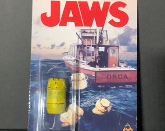 May include: A yellow and brown plastic barrel with a hook attached to the bottom. The barrel is in a plastic blister pack with a background image of a boat called "ORCA" from the movie Jaws. The text "JAWS" is in red letters at the top of the package.