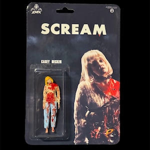 Puede incluir: Figura de acción de Casey Becker de Scream en un blíster de plástico. La figura es una miniatura de un personaje con cabello rubio, una camisa manchada de sangre y pantalones azules. El embalaje presenta la palabra "SCREAM" en letras blancas.