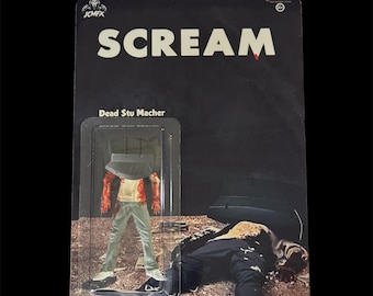 May include: A Scream action figure on a black card. The card reads "SCREAM" in white letters. The figure is of Dead Stu Macher, with a black hat, blood-stained arms, and gray pants. The figure is in a clear plastic case.