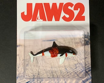 May include: A plastic package with a white background and a red "JAWS 2" logo. Inside the package is a small, white, black, and red plastic shark figure.