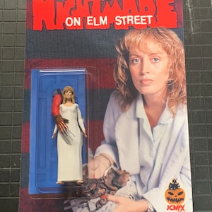 Może przedstawiać: Figurkę akcji Nightmare on Elm Street w plastikowym opakowaniu. Figurka przedstawia kobietę w białej szacie z czerwoną i brązową rękawicą z ostrzami na palcach. Tło to wzór w czerwono-niebieskie pasy z napisem "A Nightmare on Elm Street" białymi literami. Figurka trzyma nóż.