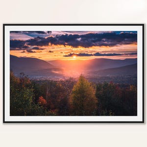 May include: Framed landscape print featuring a vibrant sunset over a valley. The sky is filled with colorful clouds, and the sun's rays illuminate the mountains and trees below. The artwork is encased in a black frame.