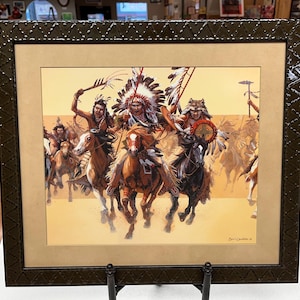 May include: Framed print depicting Native American warriors on horseback. The artwork features a dynamic scene with riders in traditional attire, including feathered headdresses and shields, set against a warm, desert-like backdrop. The frame is dark brown with a textured design.