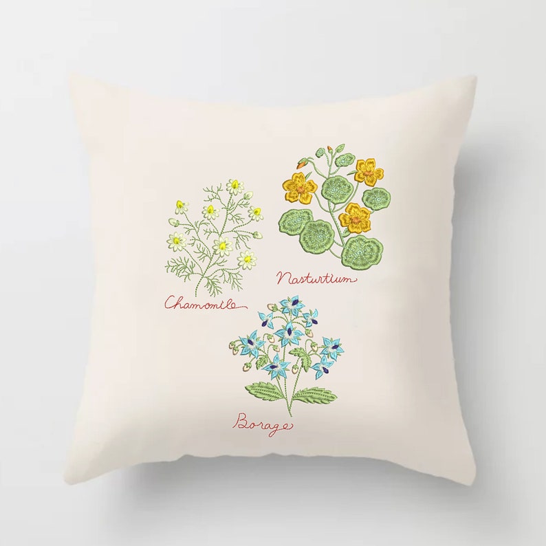 Pu&ograve; includere: Un cuscino bianco con immagini ricamate di tre fiori: camomilla, nasturzio e borragine. I fiori sono ricamati in tonalit&agrave; di verde, giallo, blu e bianco. I nomi dei fiori sono scritti in corsivo sotto ogni immagine.