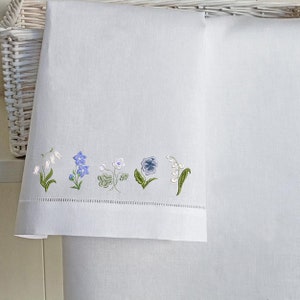 Puede incluir: Dos toallas de mano de lino blanco con un borde bordado de flores azules y blancas. Las flores incluyen una campanilla, un nomeolvides, un tr&eacute;bol, una violeta y lirio de los valles.