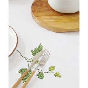 Puede incluir: Mantel blanco con hojas y enredaderas bordadas en verde. Un tenedor y un cuchillo de plata est&aacute;n colocados sobre el mantel.