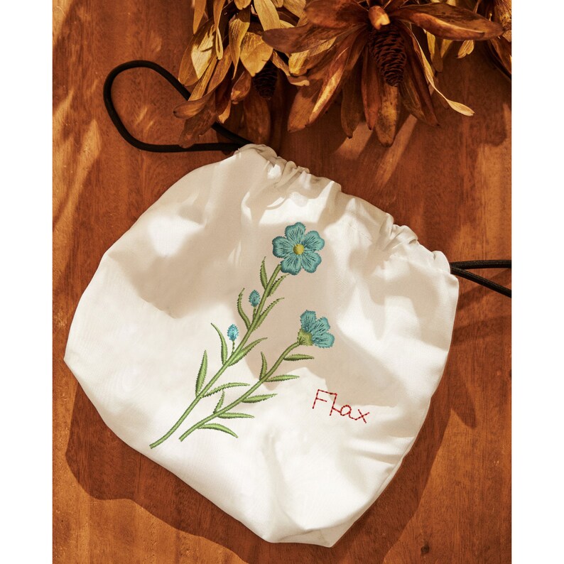 Puede incluir: Bolsa blanca de cord&oacute;n con flores azules bordadas y la palabra "Flax" en rojo.