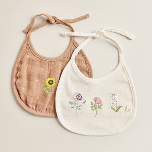 Puede incluir: Dos baberos para beb&eacute; con dise&ntilde;os florales bordados. Un babero es marr&oacute;n con un girasol y el otro es blanco con tres flores.