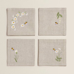 May include: Set of four linen napkins with embroidered floral and bee designs. The napkins feature a variety of flowers, including daisies, and bees.