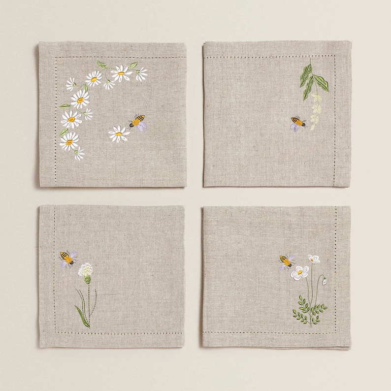 May include: Set of four linen napkins with embroidered floral and bee designs. The napkins feature a variety of flowers, including daisies, and bees.