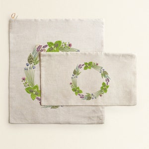 May include: Two linen fabric bags with a green and purple floral wreath design. The smaller bag is inside the larger bag.