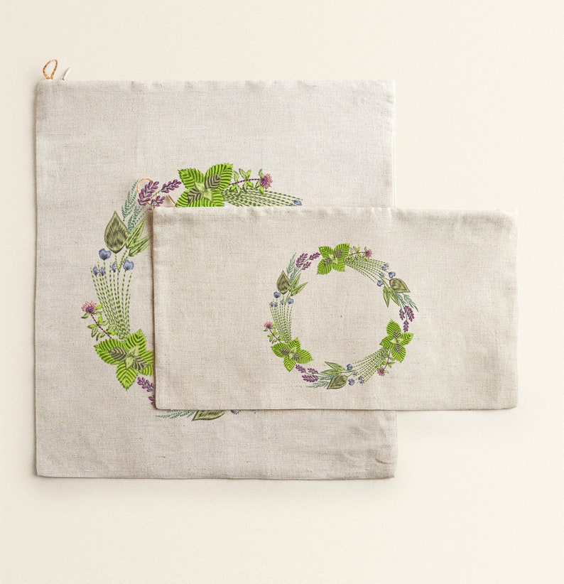May include: Two linen fabric bags with a green and purple floral wreath design. The smaller bag is inside the larger bag.