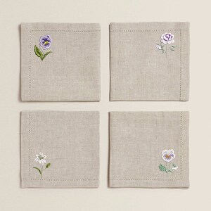 Puede incluir: Juego de cuatro servilletas de lino con dise&ntilde;os florales bordados. Cada servilleta presenta una flor diferente: una violeta, una rosa, un lirio y una violeta con un centro amarillo.