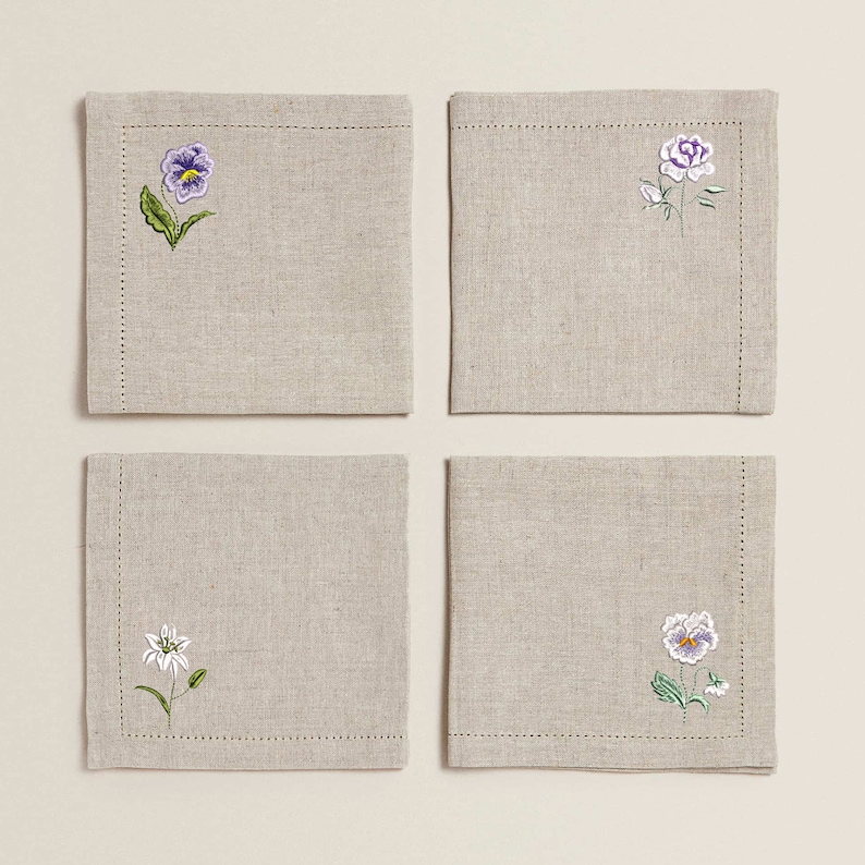 Puede incluir: Juego de cuatro servilletas de lino con dise&ntilde;os florales bordados. Cada servilleta presenta una flor diferente: una violeta, una rosa, un lirio y una violeta con un centro amarillo.