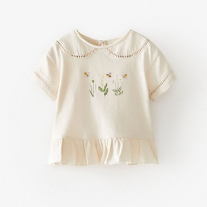 May include: A cream colored short-sleeved shirt with a ruffled hem and a peter pan collar. The shirt has a floral embroidery design with bees.