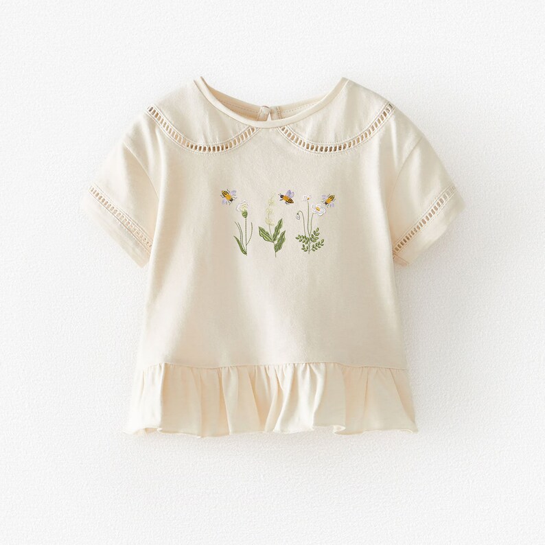 May include: A cream colored short-sleeved shirt with a ruffled hem and a peter pan collar. The shirt has a floral embroidery design with bees.